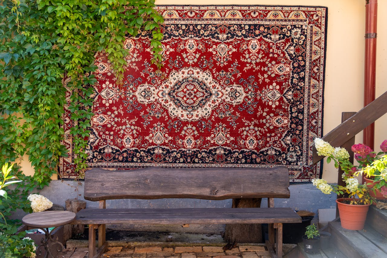 A traditional Persian rug hanging outdoors with a rustic bench, surrounded by lush greenery and potted flowers.